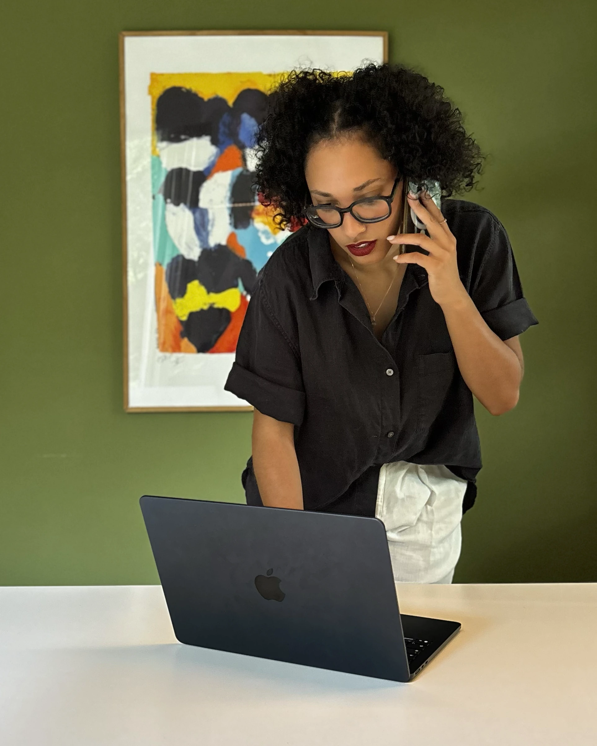 Susana standing at a desk on the phone, looking at her laptop, in front of a green wall with a painting